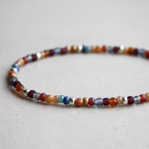 Pode incluir: Uma pulseira de contas com uma mistura de contas coloridas em tons de vermelho, laranja, azul e prata. A pulseira é enfiada em um cordão flexível e foi projetada para ser usada em volta do pulso. As contas são pequenas e uniformemente espaçadas.