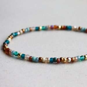 Peut inclure: Un bracelet de perles délicat composé d'un arrangement circulaire de petites perles en verre multicolores dans des tons de bleu sarcelle, or métallique, cuivre et beige translucide, formant un anneau continu, fin et circulaire.