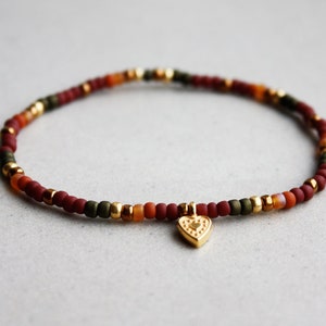 Puede incluir: Un brazalete de cuentas multicolores con un dije de corazón dorado. El brazalete tiene cuentas rojas, naranjas y verdes.