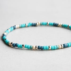 Può includere: Un braccialetto di perline con perline turchesi, blu navy e argentate. Il braccialetto è composto da piccole perline rotonde infilate insieme. Le perline sono disposte in un motivo di colori alternati. La circonferenza è di circa 17 cm.