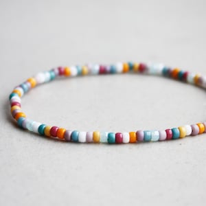 Può includere: Un braccialetto di perline colorate con piccole perline rotonde in una varietà di colori tra cui arancione, verde acqua, viola e bianco. Il braccialetto ha una circonferenza di circa 17,8 cm ed è esposto su una superficie grigia neutra.