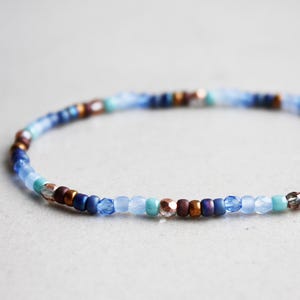Pode incluir: Uma pulseira de contas com pequenas contas azuis, castanhas e iridescentes. As contas são enfiadas para formar uma faixa flexível. A pulseira apresenta uma mistura de cores, incluindo turquesa e castanho.