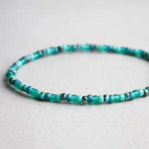 Op de afbeelding: Een delicate kralenarmband met kleine, ronde kralen in tinten van blauwgroen, turquoise en groen. De armband is aan elkaar geregen, waardoor een flexibele band ontstaat. De kralen hebben een diameter van ongeveer 0,3 cm. De armband wordt getoond op een lichtgrijze ondergrond.