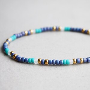 Könnte beinhalten: Ein Perlenarmband mit einem Muster aus marineblauen, türkisfarbenen, weißen und goldenen Perlen. Das Armband besteht aus kleinen, runden Perlen, die zu einem flexiblen Band aufgefädelt sind. Die Perlen sind in einem sich wiederholenden Farbmuster angeordnet.
