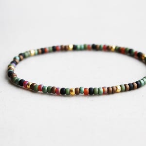 Pode incluir: Uma pulseira fina e circular de contas com um padrão repetitivo de pequenas contas de acabamento mate em tons de verde sálvia, castanho escuro, preto e vermelho suave, intercaladas com pequenas contas espaçadoras metálicas douradas.