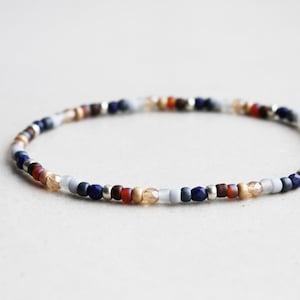 Pode incluir: Uma pulseira fina de missangas com um padrão repetitivo de pequenas contas redondas em tons de azul-marinho, vermelho-escuro, cinzento-claro e âmbar translúcido, intercaladas com pequenas contas espaçadoras de metal prateado em círculo.