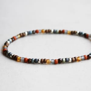Pode incluir: Uma pulseira de contas com uma mistura de pequenas contas coloridas. A pulseira inclui contas em tons de castanho, laranja, azul, cinza e prata. As contas são enfiadas para formar uma faixa flexível, adequada para o pulso.