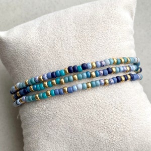 Puede incluir: Un brazalete de múltiples hilos con cuentas azules y doradas. El brazalete está hecho de pequeñas cuentas redondas en tonos azules, turquesa y dorado. Las cuentas están enhebradas en un cordón elástico transparente.