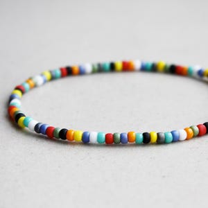 Könnte beinhalten: Ein farbenfrohes Perlenarmband mit einer Vielzahl kleiner, runder Perlen in verschiedenen Farben, darunter Rot, Gelb, Blau, Grün, Schwarz und Weiß. Das Armband ist für das Handgelenk konzipiert.