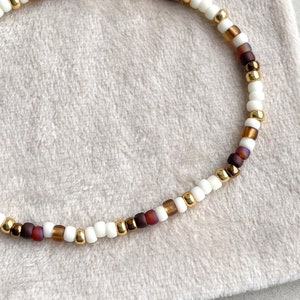 Könnte beinhalten: Ein Armband aus weißen, goldenen und braunen Perlen. Das Armband besteht aus kleinen, runden Perlen.