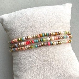 Puede incluir: Tres pulseras de cuentas en colores pastel, incluyendo rosa, verde y azul, con detalles dorados. Las pulseras están sobre un cojín gris claro, mostrando el diseño y la paleta de colores de la joyería.