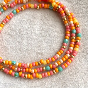 Op de afbeelding: Close-up van drie strengen kleurrijke kralenjuwelen. De kralen zijn in tinten roze, oranje, geel en turquoise, met gouden accenten. De sieraden worden gepresenteerd op een zacht, wit oppervlak.