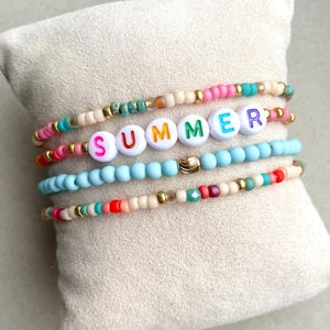 Op de afbeelding: Een verzameling armbanden met kralen met het woord "SUMMER" in kleurrijke letterkralen. De armbanden hebben verschillende kleuren, waaronder roze, blauw en goud, en worden gepresenteerd op een beige kussen.