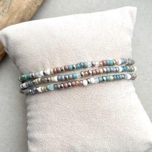 Pode incluir: Um conjunto de três pulseiras de contas com um padrão repetitivo de pequenas contas redondas em tons de azul-petróleo, branco, prata metálica e castanho suave. As contas estão enfiadas num fio flexível, criando um visual em camadas.