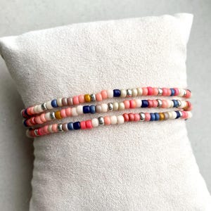 May include: Three beaded bracelets in shades of pink, white, blue, and silver. The bracelets are displayed on a white, textured surface. The small beads are evenly spaced, creating a delicate and colourful accessory.