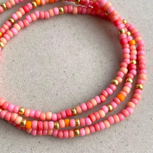 Könnte beinhalten: Nahaufnahme von drei Strängen Perlschmuck. Die Perlen sind in Rosa-, Korallen- und Orangetönen gehalten, durchsetzt mit kleinen goldfarbenen Perlen. Der Schmuck ist in einem geschichteten, kreisförmigen Muster vor einem neutralen Hintergrund angeordnet.