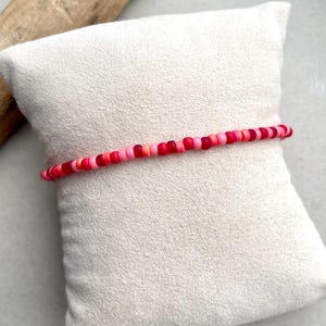 Puede incluir: Una pulsera de una sola hebra compuesta por pequeñas cuentas redondas en un degradado de colores que van desde el rosa claro y coral hasta el rojo vibrante y el carmesí intenso, dispuestas en línea continua sobre un cojín de tela cruda.