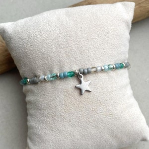 Op de afbeelding: Een delicate armband met een zilveren sterbedel. De armband is gemaakt met kleine grijze, blauwe en groene kralen.