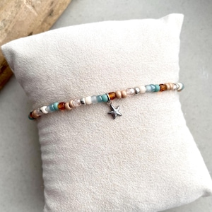 Op de afbeelding: Een kralenarmband met een reeks kleine, ronde kralen in tinten amber, turquoise, crème en metallic zilver. Een kleine, vijfpuntige zilveren sterbedel hangt aan het midden van de streng, die is samengesteld uit verschillende texturen.