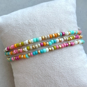 Może przedstawiać: Wielokolorowe bransoletki z koralików ze złotymi akcentami. Bransoletki są ułożone na białej powierzchni.