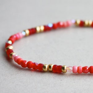 Könnte beinhalten: Ein Perlenarmband mit roten, rosa und goldenen Perlen. Das Armband besteht aus kleinen, runden roten Perlen, die mit rosa und goldfarbenen Perlen durchsetzt sind. Das Armband ist flexibel und für das Handgelenk konzipiert.