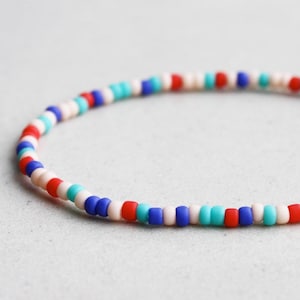May include: A beaded bracelet featuring small, round beads in a repeating pattern of red, white, blue, and turquoise. The bracelet is curved, suggesting it is flexible and designed to be worn on the wrist.