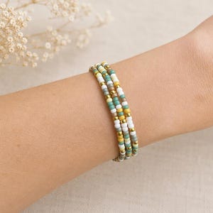 May include: A multi-strand bracelet composed of three rows of small seed beads in a colour palette of white, mustard yellow, sage green, grey, and metallic gold. The beads are arranged in a repeating pattern, creating a layered appearance on the wrist.