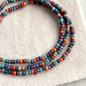 Può includere: Tre fili di collane di perline colorate. Le perline sono piccole e cilindriche, nelle tonalità del blu, arancione, rosso e viola. Le collane sono disposte in un design a strati, con uno sfondo morbido e neutro.