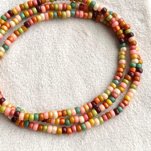 Könnte beinhalten: Drei Stränge farbigen Perlenschmucks. Die Perlen sind klein und rund, in den Farbtönen Rosa, Orange, Grün, Gelb und Braun. Der Schmuck ist kreisförmig auf weißem Hintergrund angeordnet.