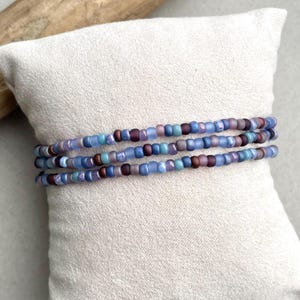 Peut inclure: Trois bracelets de perles aux tons de bleu, violet et marron sont présentés sur un coussin blanc. Les bracelets sont composés de petites perles rondes, créant un accessoire délicat et coloré. Les perles ont des finitions mates et translucides.