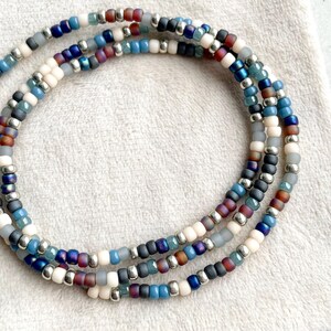 Pode incluir: Três pulseiras de contas em tons de azul, branco, cinza e castanho com contas prateadas. As pulseiras estão dispostas em um padrão circular sobre uma superfície branca e texturizada. As contas são pequenas e espaçadas uniformemente.
