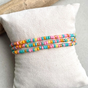 Könnte beinhalten: Drei Perlenarmbänder in Pastellfarben Rosa, Gelb, Türkis und Orange. Die Armbänder bestehen aus kleinen, runden Perlen und sind auf einem weißen Kissen angeordnet. Die Perlen haben eine glänzende Oberfläche und sind aufgefädelt.
