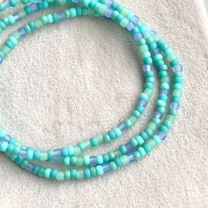 Op de afbeelding: Drie strengen kralen sieraden in tinten turquoise, lichtblauw en zeegroen. De kralen zijn klein en vierkant en liggen op een witte, getextureerde ondergrond.