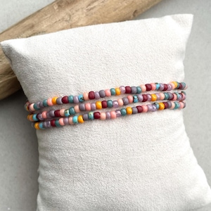 Op de afbeelding: Drie kralenarmbanden in roze, paarse, turquoise, oranje en bordeaux tinten worden getoond op een wit kussen. De armbanden zijn gemaakt van kleine, kleurrijke kralen en zijn om elkaar heen gewikkeld.