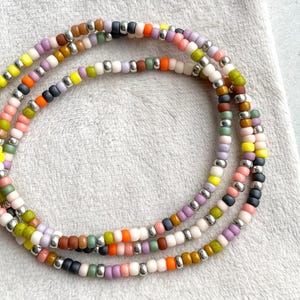 Pode incluir: Três pulseiras de contas com uma variedade de cores, incluindo amarelo, rosa, laranja, verde, roxo, castanho e preto. As contas são pequenas e redondas, com espaçadores de metal prateado. As pulseiras estão dispostas em um padrão circular.