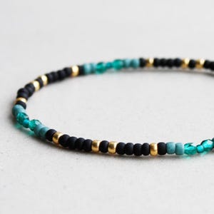 Könnte beinhalten: Ein zartes Armband aus schwarzen, goldenen und türkisfarbenen Perlen. Das Armband besteht aus kleinen, runden Perlen und hat ein schlichtes, elegantes Design.