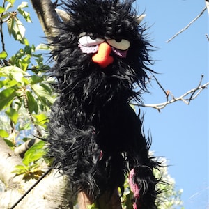 May include: A black furry puppet with a large orange nose and purple eyelids. The puppet is perched on a tree branch.