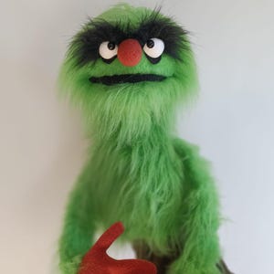 Könnte beinhalten: Eine handgefertigte grüne Marionette mit pelziger Textur. Sie hat schwarze Augenbrauen, einen schwarzen Mund, eine rote Nase und weiße Augen. Die Hände der Marionette sind rot. Die Marionette ist ein skurriles, weiches Spielzeug.