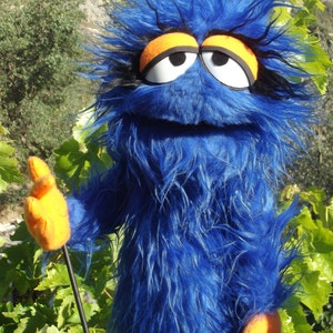 May include: Blue fuzzy hand puppet with orange and white eyes and a black mouth. The puppet is wearing orange gloves and is holding a black stick.