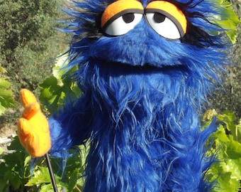 May include: Blue fuzzy hand puppet with orange and white eyes and a black mouth. The puppet is wearing orange gloves and is holding a black stick.