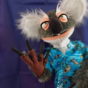 May include: A koala puppet with a gray and white fur head, wearing a blue Hawaiian shirt with a dolphin print. The puppet has green eyes, a black nose, and is making a rock-and-roll hand gesture.