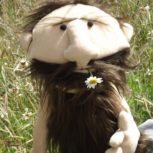 Op de afbeelding: Een bruine pop met een lange, borstelige baard en een witte bloem in zijn mond. De pop heeft een grote, ronde neus en een serieuze uitdrukking.
