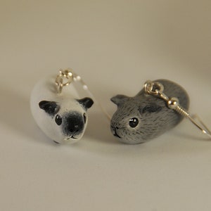 May include: A pair of dangle earrings featuring sculpted guinea pig charms. One charm is white with a black nose and ears, while the other is solid grey with detailed fur texture. Both charms are attached to silver-toned metal earring hooks.
