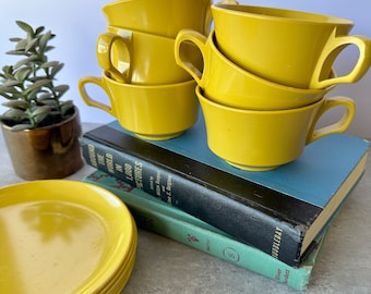 May include: A set of six yellow ceramic teacups and saucers. The cups and saucers are stacked on top of each other. The cups have a rounded shape and a handle on each side. The saucers are flat and have a slightly raised edge.