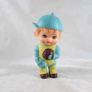 May include: A blue and yellow plastic toy baseball player with a blue cap. The toy is holding a brown baseball.