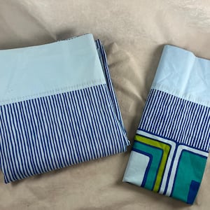 May include: A folded flat sheet and a pillowcase featuring a light blue top border and a lower section with vertical blue stripes. The pillowcase also displays a geometric pattern in shades of green, teal, and blue along the bottom edge.