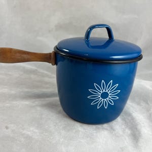May include: A blue enamel saucepan with a lid and wooden handle. The pot features a white floral design on the side. The handle is made of light brown wood. The pot is designed for cooking.