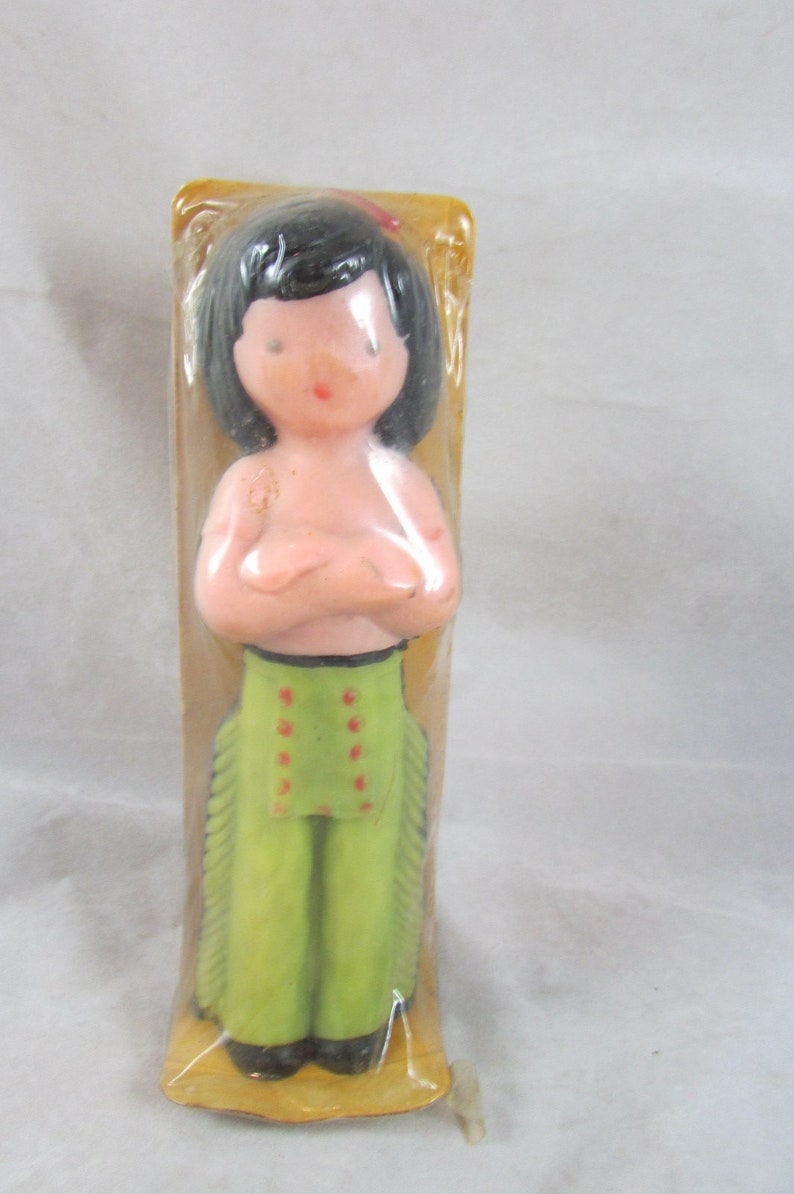 May include: A plastic doll figurine of a Native American person wearing a green and red outfit with fringe. The doll is packaged in clear plastic.