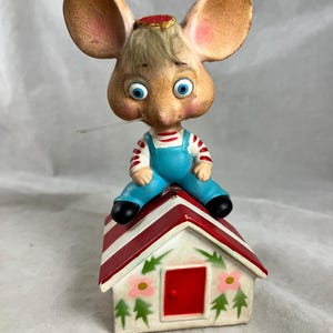 May include: A vintage mouse figurine with large ears, blue eyes, and a red and white striped shirt and blue overalls. The mouse sits atop a small house with a red and white striped roof and floral details.