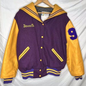 May include: Purple and gold varsity jacket with leather sleeves. The jacket has a sailor collar, a button closure, and a number 9 on the right sleeve. The name "Barnette" is embroidered on the left side of the jacket.